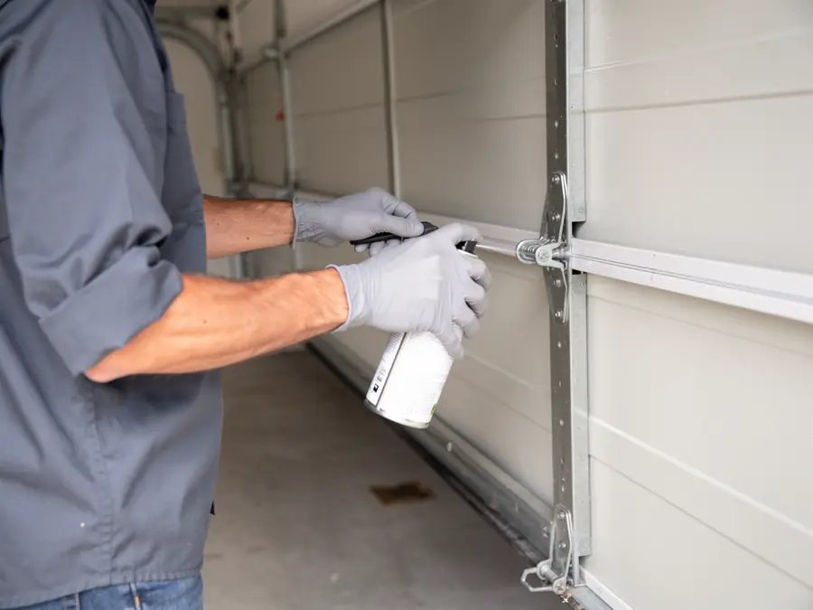 A technician in gloves sprays lubricant on a garage door hinge.